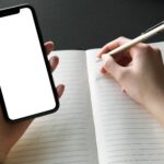 Person writing in a notebook while holding a smartphone with a blank screen, ideal for mockups.