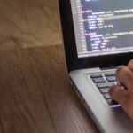 A person is typing code on a laptop, focusing on the screen with programming script.